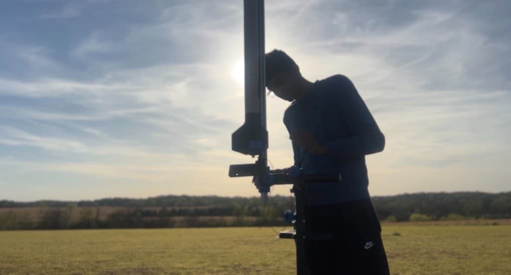 Student preparing a model rocket on a vertical launch rail in an open field at sunset